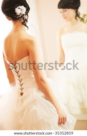 Wedding shot of a slim beautiful woman wearing luxurious wedding dress standing opposite to a mirror.