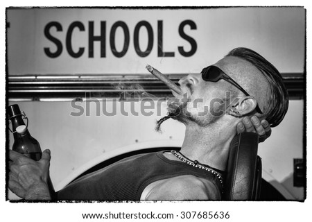 Bad Boys do not like school: Tough guy with sparrow beard, undercut and blue jeans sitting on his chopper bike in front of a yellow, American school bus. He is smoking cigar and drinking beer.