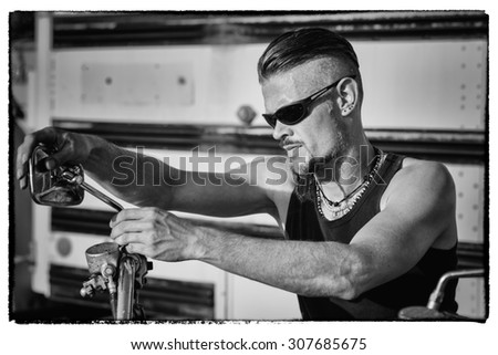 Tough guy with sparrow beard, undercut, black rip shirt and sun glasses lolling on his chopper motorcycle in front of a yellow, American school bus.