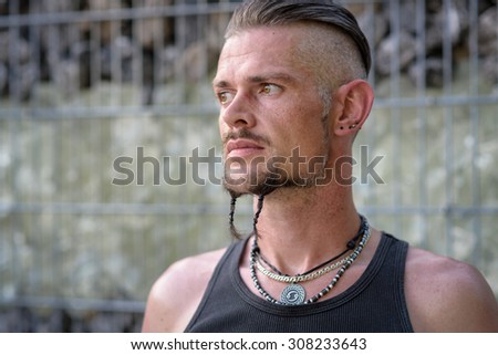 Tough guy with sparrow beard, undercut and black rip shirt standing thoughtful in front of a wall of stones