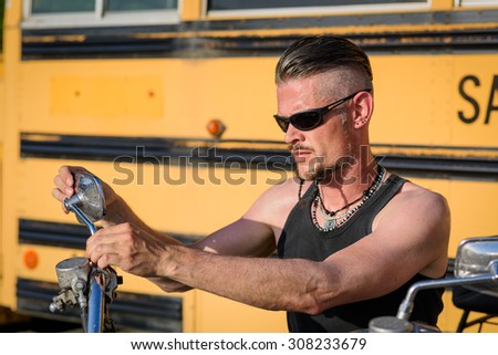 Tough guy with sparrow beard, undercut, black rip shirt and sun glasses lolling on his chopper motorcycle in front of a yellow, American school bus.