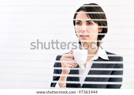 Business woman with cup of coffee looking through a venetian blind in an office