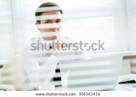 Handsome young businessman working at laptop in office. View through blinds