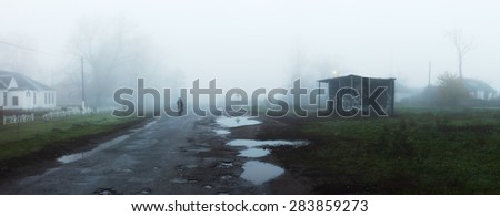 Rural landscape with road and bus stop in fog. Silhouette of man walking on misty village road. Loneliness, nostalgia, sad mood