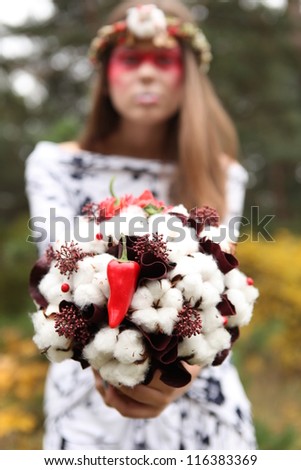 priestess of Samhain in a wreath and with a bright red makeup extends bouquet of cotton and peppers