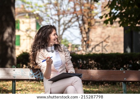 happy young woman caucasian asian in pastel pink outfit sitting on a bench laughing and writing in the park