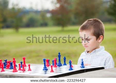 Elegant young boy in white shirt learning to play chess with blue and red chess pieces on wood table in the park