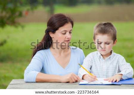 Little young boy in white shirt and his mother write or draw with a pencil on a sheet of paper on wood table in the park