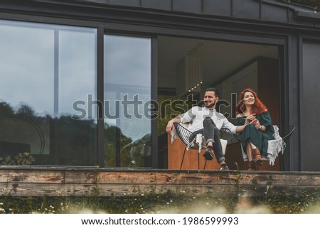 Front view of happy family sitting outdoor on the terrace of country house. Stylish young couple on weekend sits and enjoys the nature view on the terrace of their modern home with panoramic windows.