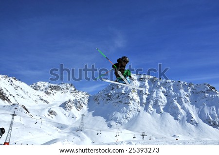 skier in green and black clothes performing a very high jump against a backdrop of snow covered mountains and ski ilfts