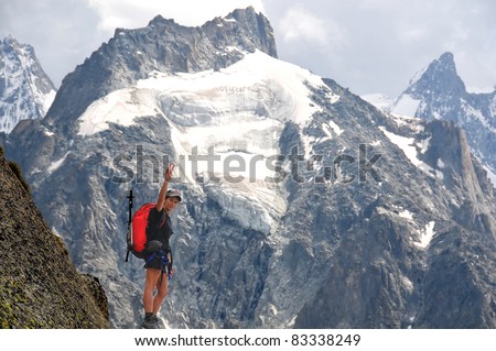 a woman climber reaches the summit of a high mountain and makes a victory sign, with other  ice covered peaks in the background