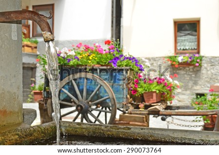 A traditional water spout filling a trough with a flower filled wagon in the background. Flowers need plenty of watering
