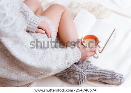 Soft photo of a woman in a cozy sweater on the bed with the old book and a cup of tea with milk in their hands