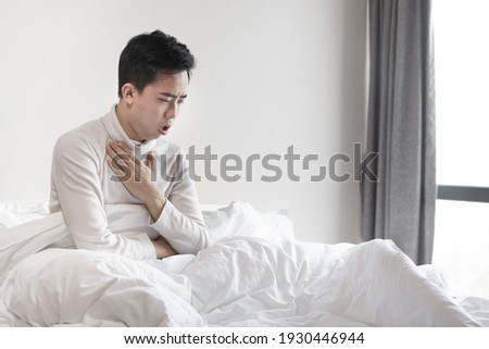 Man sitting on the bed is coughing with hand covered his chest 