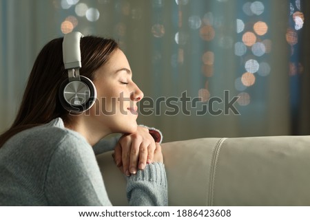 Profile of a woman with closed eyes feeling and listening to music with wireless headphones sitting on a couch in the night at home