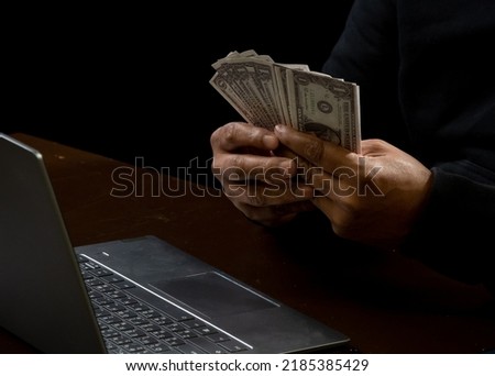 Computer and hands of a man wearing a black shirt, sitting on a chair and a table, is a thief, holding money, counting the amount obtained from hijacking or robbing, in a pitch-black room. Foto stock © 