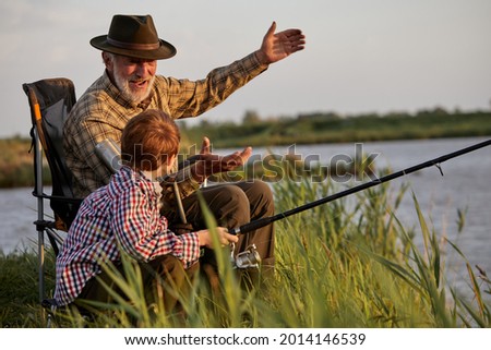 Grandfather and kid boy together fishing in the evening at sunset time in summer day on river in countryside, side view on caucasian multi-generation family, elderly man teach child boy to fish