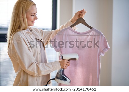 Woman using steaming iron to ironing casual t-shirt at home. Girl doing stream vapor iron for press clothes in hand. Blonde female doing household chores, housewife using smart device for ironing