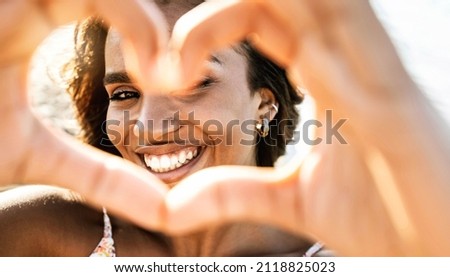 Photo of Close up image of smiling woman in swimwear on the beach making a heart shape with hands - Pretty joyful hispanic woman laughing at camera outside - Healthy lifestyle, self love and body care concept