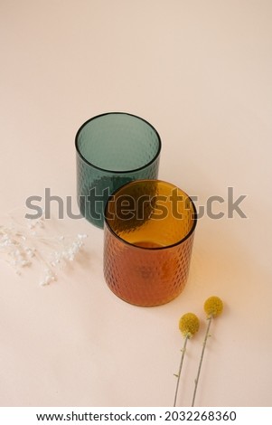 Glass orange and green cups on pink background with decoration. Emerald green cup. Amber orange cup. Couple glass cups with flowers. Tea cups. Coffee cups. Smoothies cup. Water cup.