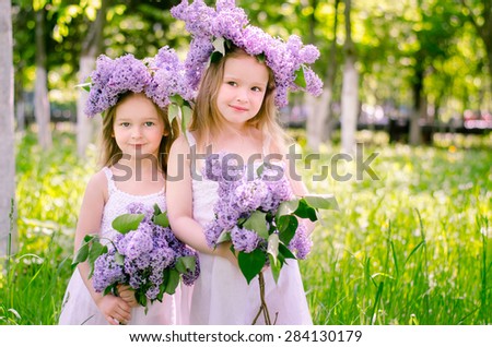 Young beautiful blond girls dressed in a white dress with a bouquet of lilac in the hands and wreaths of purple flowers on their heads are in the park on a sunny morning and smile.