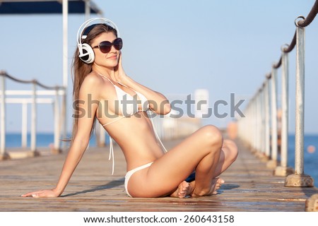 young woman in headphones sitting on beach and enjoying beautiful sunset over the sea, rear view
