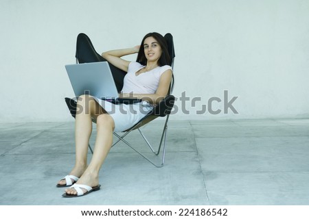 Young woman sitting back in chair, using laptop, full length portrait