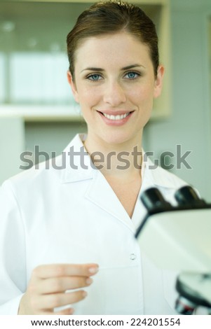 Female lab worker with microscope, holding up slide, smiling at camera, portrait