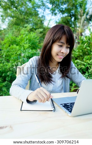 happy asian young female taking note on table with laptop computer in the garden. People, technology and education concept