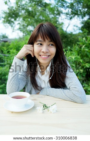Female student girl outside in garden listening to music on earphones with cup of tea and flowers on wooden table.