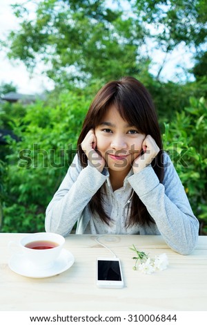 Female student girl outside in garden listening to music on earphones with cup of tea and flowers on wooden table.