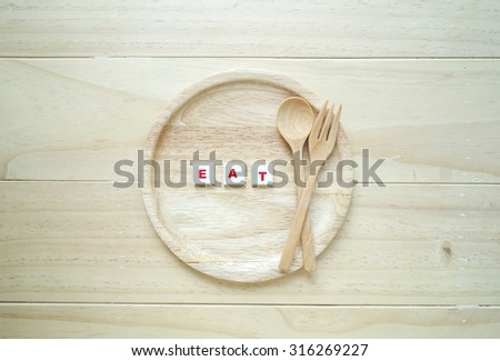 Wooden plate with hand holding spoon and fork on wooden table. The word EAT spelled using word tiles