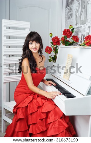 Young beautiful woman in red evening dress sits at the piano. Looking at the camera