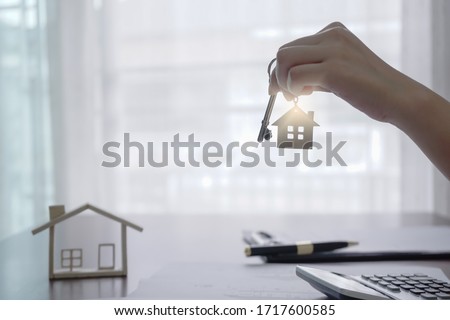 Real estate agent holding house key on house shaped keyring on table with house designs document, calculator, model house. Concept of Investment property.
