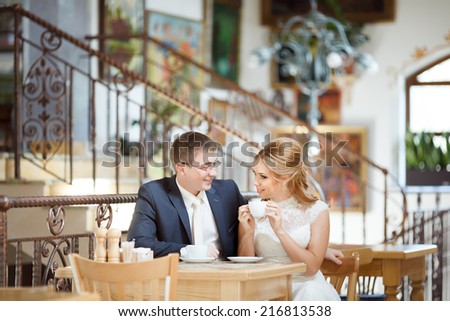 Two beautiful young people, bride and groom sitting at a table in a cafe.With a good mood, they are celebrating their wedding day, drinking coffee from a white cup.
