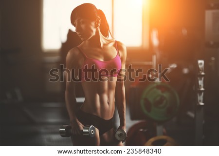Brutal athletic woman pumping up muscles with dumbbells. Brunette sexy fitness girl in pink sport wear with perfect body in the gym posing before training set. Attractive fitness woman