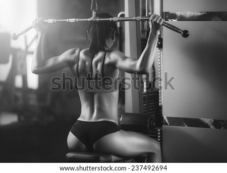 Black and white photo of a strong woman from behind. Brunette sexy fitness girl in sport wear with perfect body in the gym posing before training set. Attractive fitness woman, trained female body