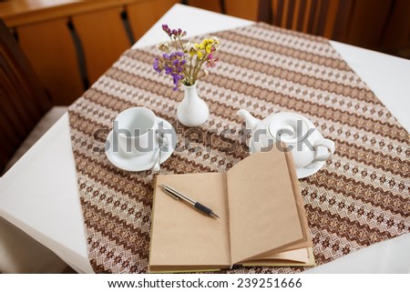 Ballpoint pen and notebook on the table in a cafe, standing next to a cup of tea, teapot and vase with a bouquet of flowers, top view