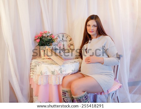 Pregnant woman reading a book while sitting at a table on which stands a vase with pink flowers and watches