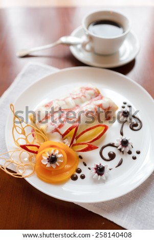 Pancakes with cherry jam beautifully decorated with flowers of fruit and chocolate on a white plate, which stands on a wooden table near the window in the interior cozy cafe. Shallow depth of field.