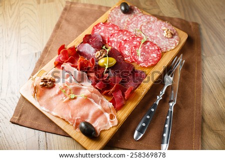 Variety of meats, sausages, salami, ham, olives, laid out on a wooden board close-up, horizontal, are next to a knife and fork