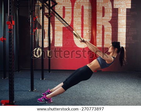 Workout on rings. Athletic young brunette woman in gray sportswear, trains on the rings in the gym with red wall on which is written the word \