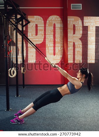 Workout on rings. Athletic young brunette woman in gray sportswear, trains on the rings in the gym with red wall on which is written the word \