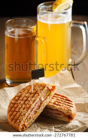 One burger with ham, onion, cucumber and cheese on a wooden plate sprinkled with sesame seeds, paper napkins, small black flag and two glasses of beer on a brown wooden background, close-up