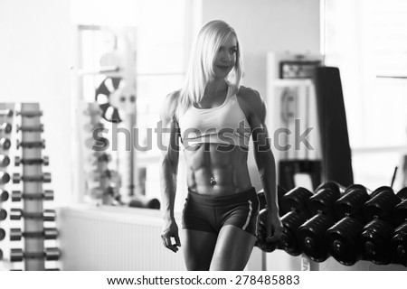 Black and white photo of a strong woman. Young muscular woman bodybuilder with white hair and tanned body posing in white gym at the counter with dumbbells