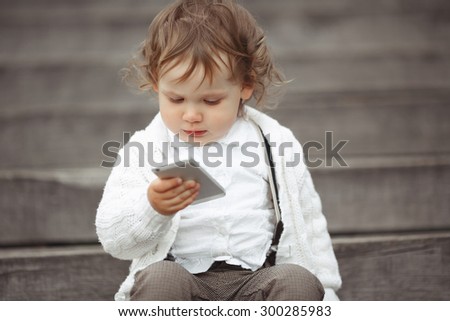 Cute little girl in white knitted sweater playing with mobile phone outdoors