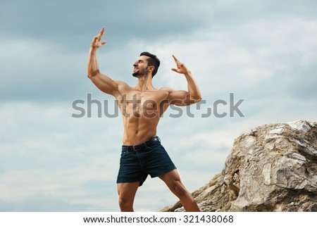 Muscular male torso on a background sky. Athletic and muscular man with naked torso stands on top of a mountain. Handsome young guy posing sporty appearance on the sky background, looking up