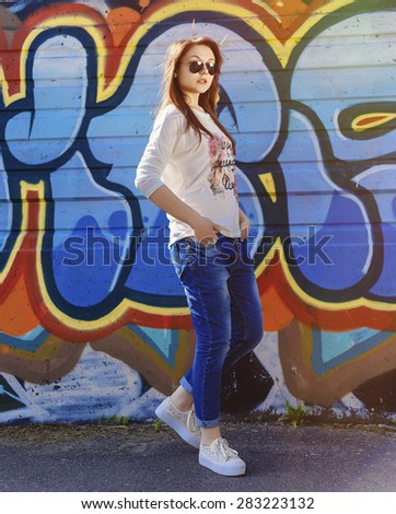Full Length Portrait of Trendy Hipster Girl Standing at the Graffiti Brick Wall Background. Urban Fashion Concept. Copy Space.