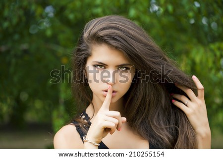 Young beautiful brunette woman has put forefinger to lips as sign of silence, against green summer garden