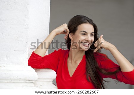 Beautiful woman making a call me gesture, happy girl in red dress on white wall background